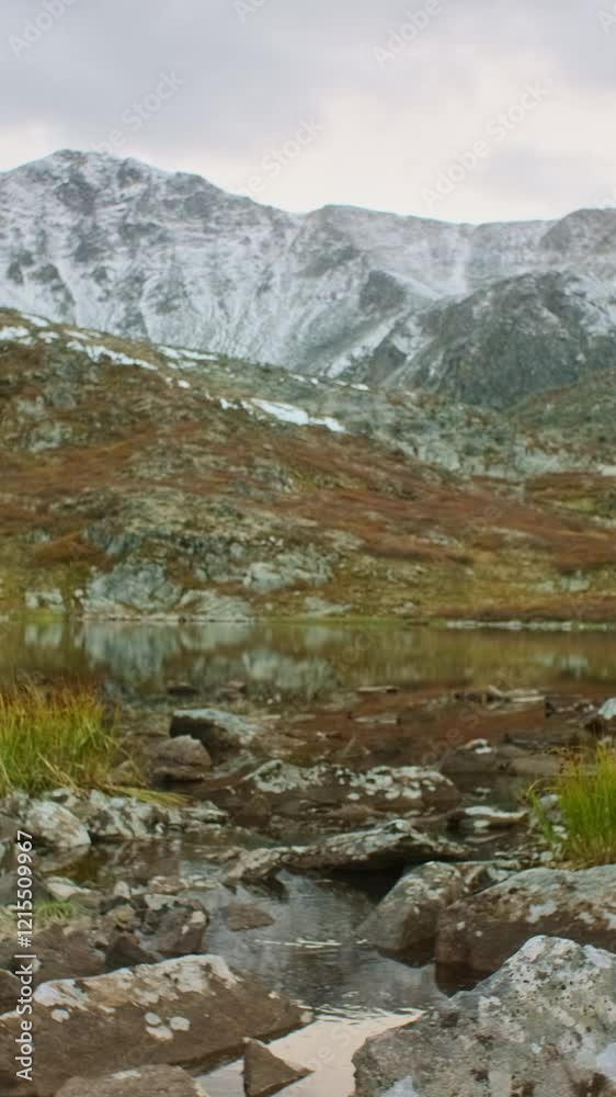 Female hiker with backpack carefully crossing stream on rocks near mountain lake with snowy peaks in the background, enjoying a challenging hike in autumn nature Female hiker with backpack carefully crossing stream on rocks near mountain lake with snowy peaks in the background, enjoying a challenging hike in autumn nature