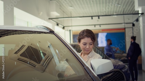 Customer in showroom looking for new vehicle to purchase or lease, running hand on car exterior to check quality. Woman evaluating automobiles in dealership before buying, camera B photo