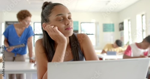 In high school, teenage girl daydreaming at desk with teacher and students in classroom