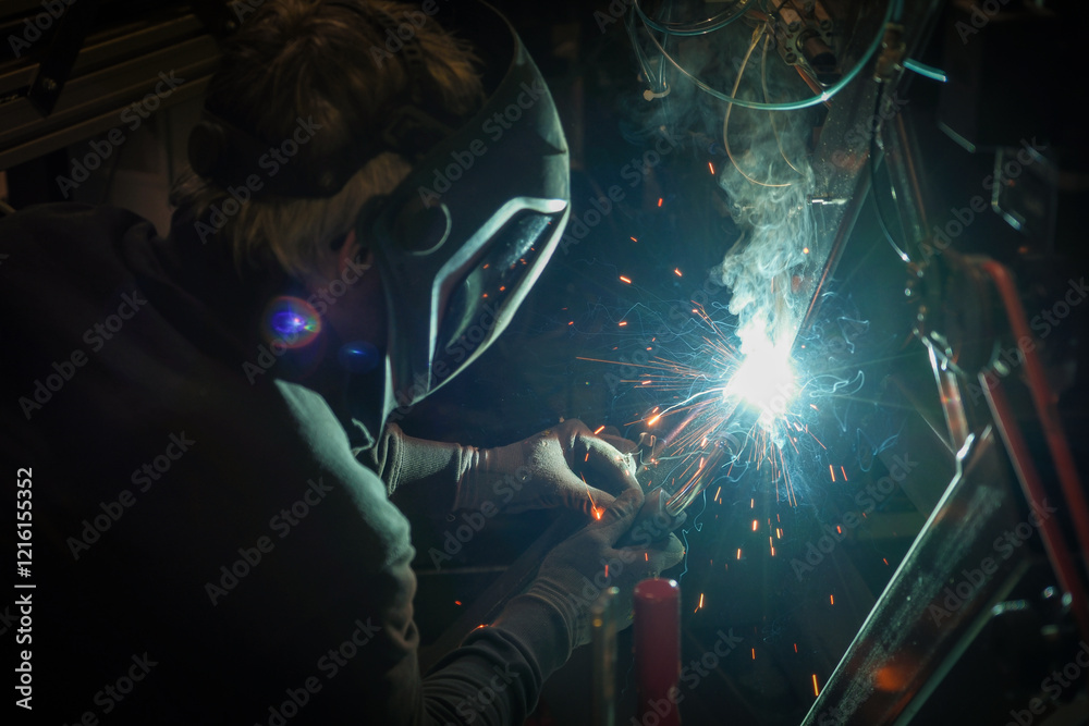 a person wearing a welding mask works on metal with a welding torch, creating sparks through the welding process
