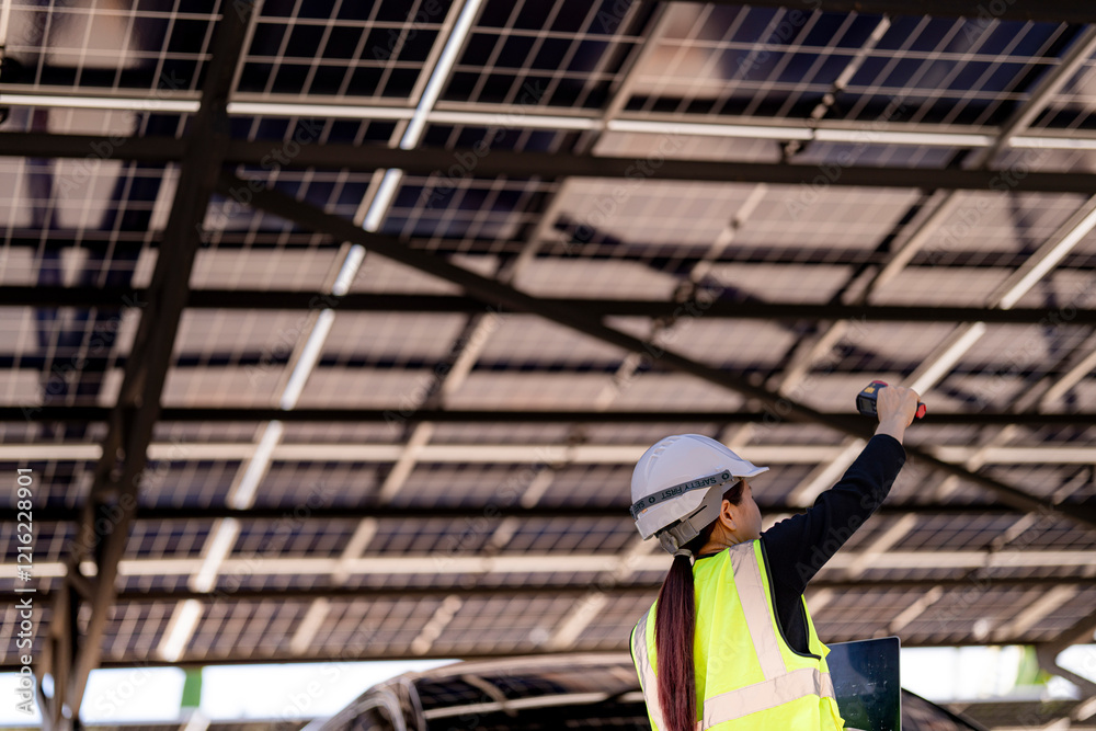 Fototapeta premium A female technician in a hard hat and reflective vest conducts a solar panel inspection with a laptop and a handheld device under a solar carport structure. The panels and sunlight frame the scene.