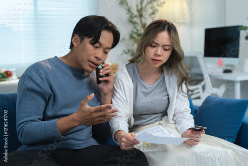 Young couple feeling worried about their financial problems, holding bills and credit card while making a phone call to the bank, sitting on sofa at home