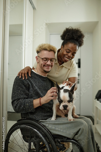 Vertical shot of cheerful man using wheelchair petting cute French bulldog sitting on lap while African American girlfriend giving him heartfelt embrace at home photo