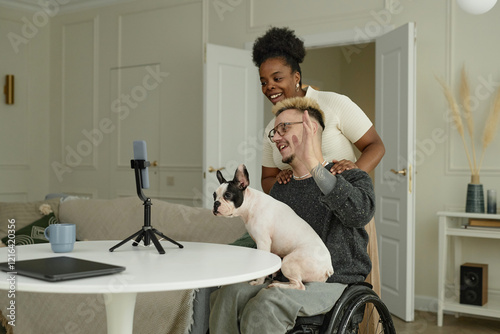 Candid shot of happy man using wheelchair accompanied by girlfriend and cute French bulldog posing together for photo on phone mounted on tripod, while having good time at home photo