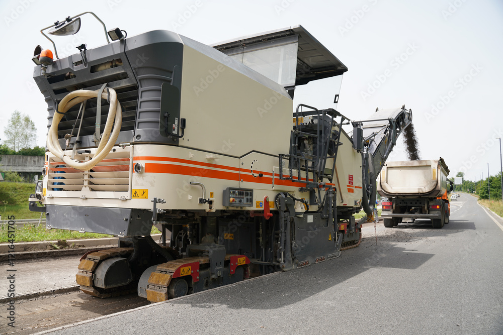 An asphalt milling machine operating on a highway construction site, milling asphalt and dumping the milled material into the cargo area of a dump truck.