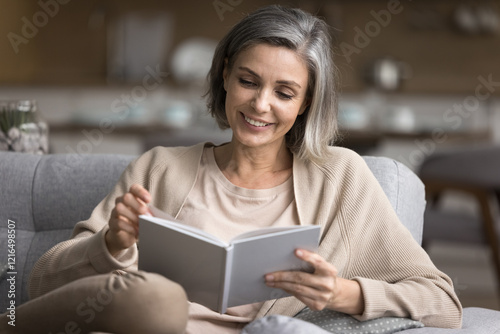 Smiling 50s female sit on sofa read book engaged in self-study using paper textbook to get knowledge spend evening at home for pleasant hobby discover fantasy world with bestseller by favorite author