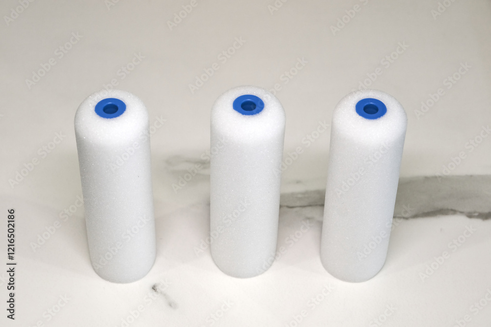 White foam paint roller refills with blue plastic cores, standing upright on a neutral background. Ideal for smooth surface painting. White foam paint roller refills with blue plastic cores, standing upright on a neutral background. Ideal for smooth surface painting.