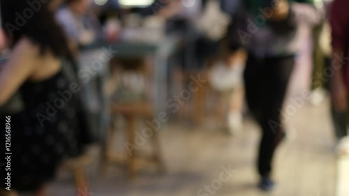 A blurry image of a restaurant with people sitting at tables and a man walking by. Scene is casual and relaxed, as people are enjoying their time at the restaurant