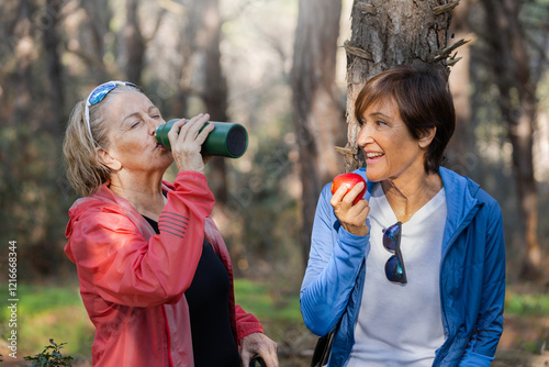 Two elderly women pausing to rehydrate during a trek in the woods. Elderly woman drinks from bottle with eyes closed while a friend eats an apple. Women refreshment on the nature. Horizontal