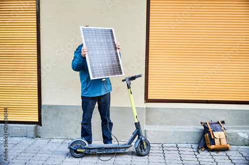 Male person holds photovoltaic solar panel, for charging electric scooter in urban settings. Integration of solar power as sustainable energy source, promoting eco-friendly urban transportation. photo