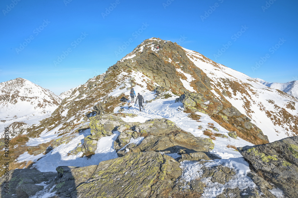 SHikers with backpacks trek along a snowy mountain trail under a brilliant blue sky, showcasing the adventurous spirit and beauty of winter mountain landscapes