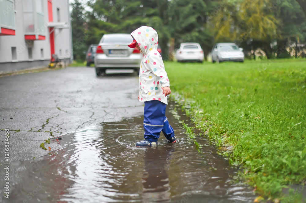 A child in a vibrant polka-dot raincoat plays joyfully in a puddle on a rainy day, while cars are parked in the background. The image captures a playful and carefree moment