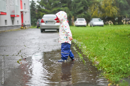 Obraz na płótnie A child in a vibrant polka-dot raincoat plays joyfully in a puddle on a rainy day, while cars are parked in the background. The image captures a playful and carefree moment w salonie
