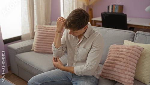Cuadro en lienzo Young man in a living room clutching his elbow in pain while sitting on a couch in a home environment with light purple walls and comfortable furnishings