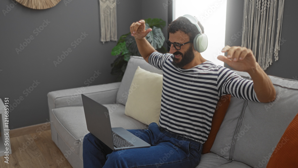 Fototapeta premium Young man with a beard enjoying music through headphones while sitting on a couch with a laptop in a cozy living room.