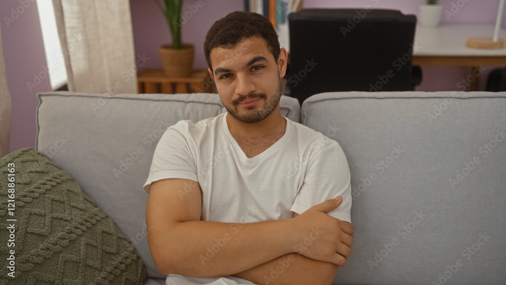 Young hispanic man sitting relaxed on a couch in the living room of a house with arms crossed and a neutral expression Young hispanic man sitting relaxed on a couch in the living room of a house with arms crossed and a neutral expression