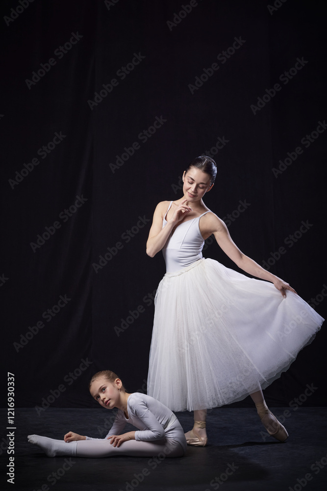 Adult ballerina standing gracefully adjusting her tutu, young ballerina lying in stretch position, both dressed in white against black studio background. Concept of ballet, elegance and beauty. Ad Adult ballerina standing gracefully adjusting her tutu, young ballerina lying in stretch position, both dressed in white against black studio background. Concept of ballet, elegance and beauty. Ad