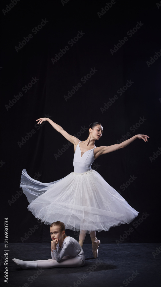 Fototapeta premium Adult ballerina spinning elegantly in tutu while young ballerina poses on floor thoughtfully both dressed in white, against black studio background. Concept of ballet, elegance and beauty, grace. Ad