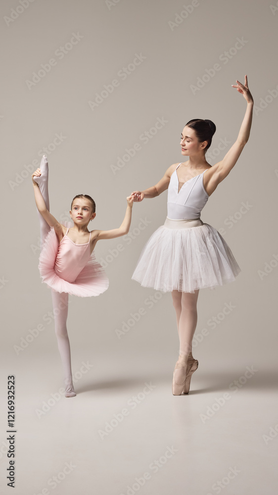 Naklejka premium Ballerina in white tutu supports little girl, dancer in pink tutu performing leg stretch, emphasizing guidance and flexibility against white background. Concept of ballet, elegance and beauty, grace.