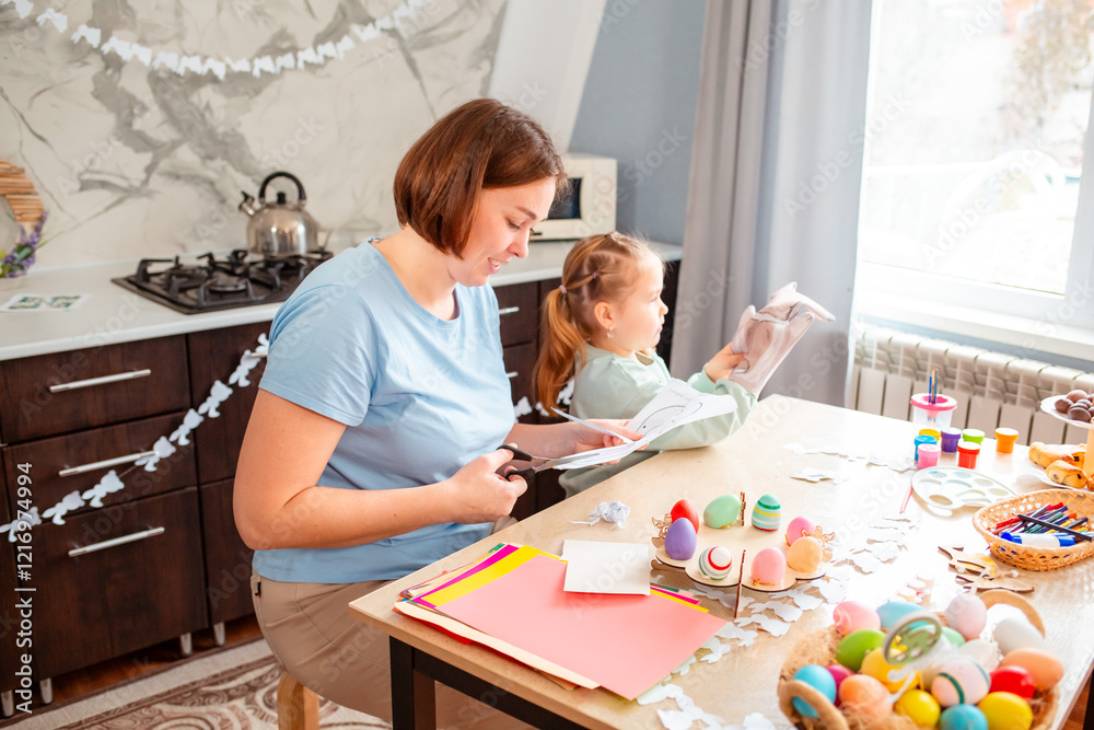 Side view of young Caucasian mother and her daughter carve a bunny mask for the Easter spring holiday. The family is sitting at the kitchen table and making festive decorations Side view of young Caucasian mother and her daughter carve a bunny mask for the Easter spring holiday. The family is sitting at the kitchen table and making festive decorations