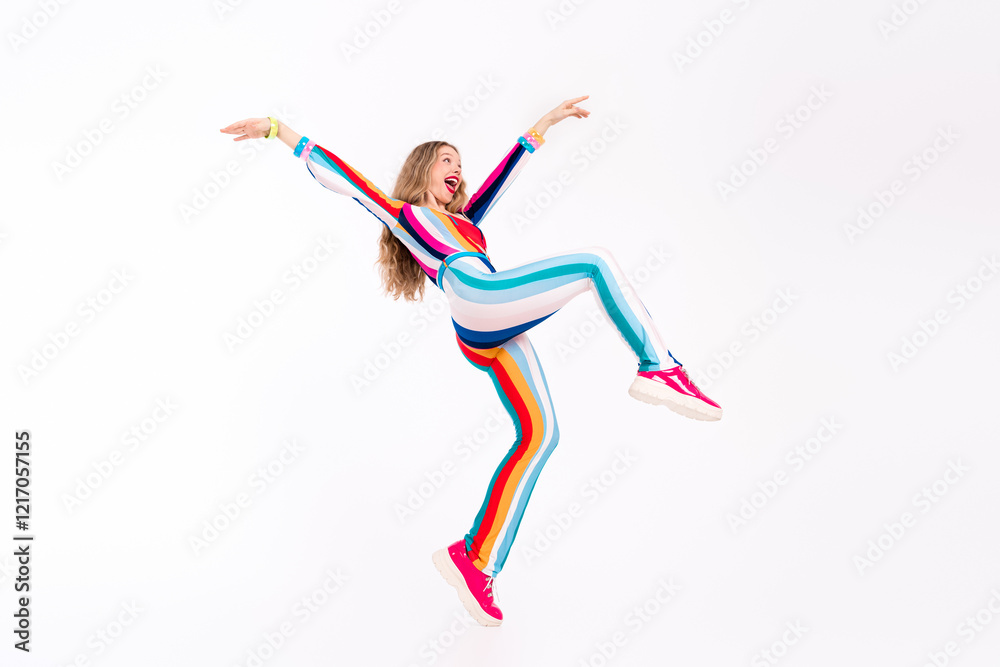 Fototapeta premium A young woman with long blond hair joyfully dancing in a vibrant striped jumpsuit against a white background