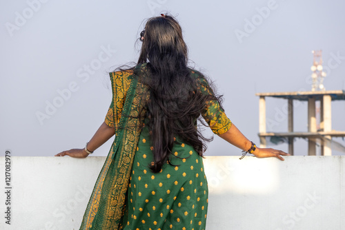 A Bengali woman, wearing a green block-printed salwar kameez and with her long hair down, stands alone on the rooftop, looking up at the sky in the afternoon. Back view. photo