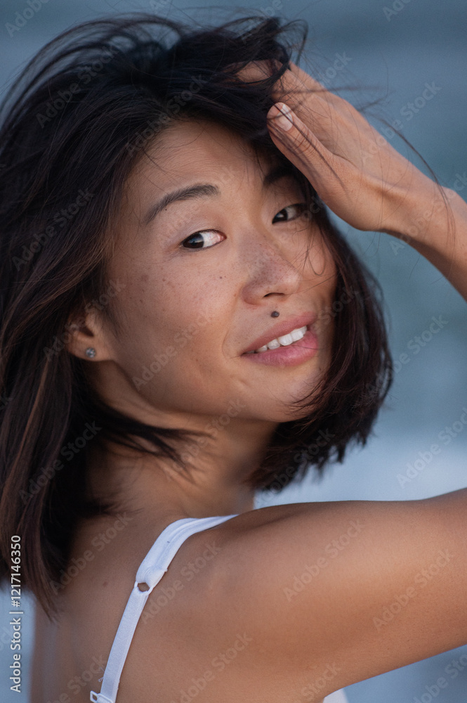 Fototapeta premium close-up portrait of an Asian woman on the beach, gently fixing her hair with her hands, looking into the camera, and smiling. Her natural beauty, freckles, and beauty mark above her lip are highlight