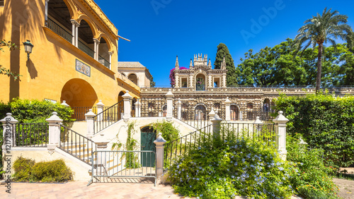 The Alcazar of Seville, historic royal palace in Seville town, Andalusia, Spain. Spanish architecture and gardens on a sunny day. Stone buildings, stairs, and greenery create a picturesque scene.