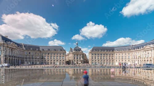 Place de la Bourse and Miroir d'eau hyperlapse in Bordeaux, France, reflecting historic architecture. Tourists enjoy the landmark and the fountain. Timelapse of traffic and tram passing by.