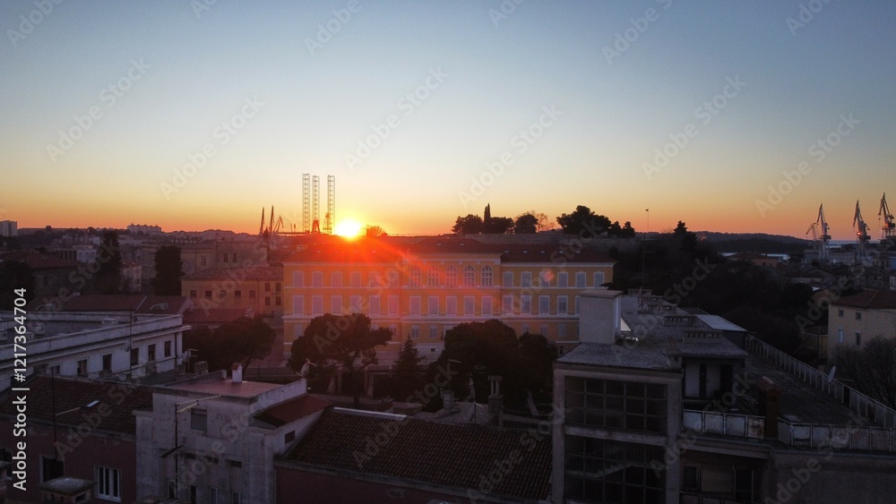 Fototapeta premium A stunning aerial view of Pula during sunset, capturing the warm, golden hues of the sky as they blend with the vibrant blue of the Adriatic Sea. This beautiful landscape offers.