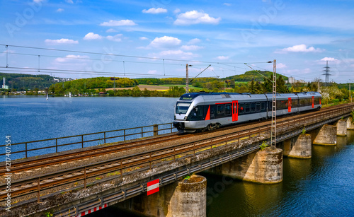A modern electric railcar crosses the Harkortsee, a reservoir of the Ruhr river, near Wetter on the route from Hagen to Dortmund. Landscape panorama with old railroad bridge and water sports area.