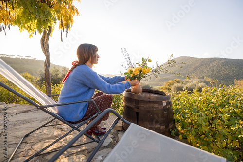 Slika na platnu A woman arranging yellow flowers in a vase on a rustic barrel table, surrounded by the serene beauty of the Tuscan countryside