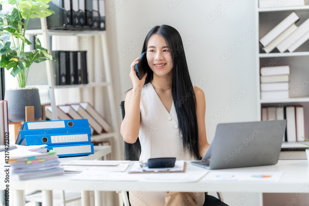 Fototapeta premium Confidently Connecting: A young Asian businesswoman smiles brightly as she conducts a phone call, demonstrating professionalism and efficiency at her desk with a laptop and documents. 