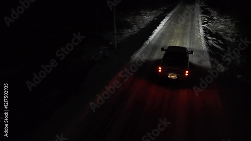 Aerial shots of a Pickup truck is driving down a snowy road at night. The headlights are on, illuminating the road ahead.