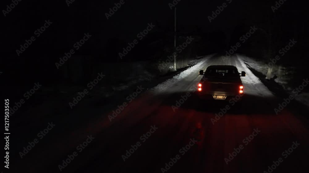 Aerial shots of a Pickup truck is driving down a snowy road at night. The headlights are on, illuminating the road ahead.