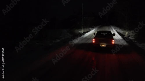 Aerial shots of a Pickup truck is driving down a snowy road at night. The headlights are on, illuminating the road ahead.