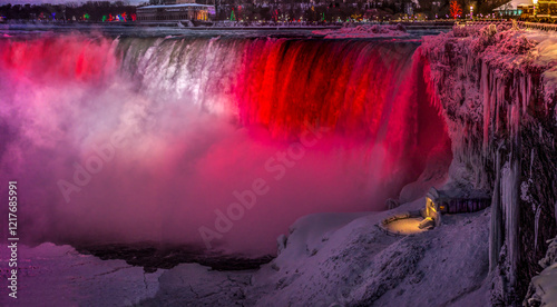 Fototapeta Naklejka Na Ścianę i Meble -  A stunning view of Niagara Falls is illuminated in the Canadian Flag colours, red and white,  at night during winter with ice formations and a glowing lookout point.