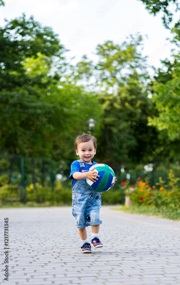 Naklejka premium Child plays ball outside. A joyful boy runs on a pathway holding a colorful ball in a park surrounded by greenery and flowers.