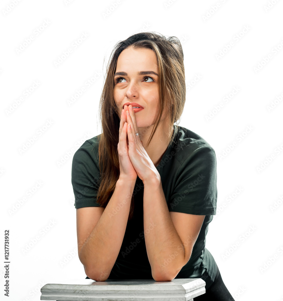 Thoughtful young woman, hands clasped. A young woman sits on a stool with her hands clasped, gazing upwards thoughtfully against a bright background. Thoughtful young woman, hands clasped. A young woman sits on a stool with her hands clasped, gazing upwards thoughtfully against a bright background.