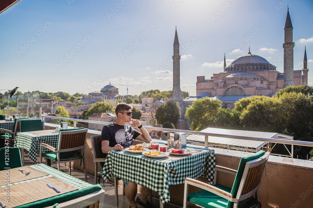 Naklejka premium Young man enjoying breakfast on rooftop terrace with stunning view of Hagia Sophia in Istanbul, Turkey on sunny day