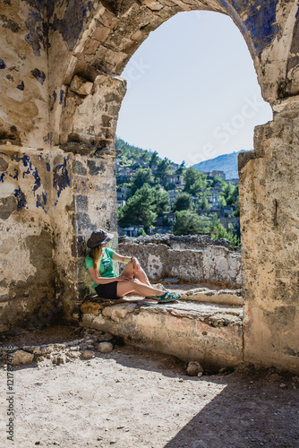 Woman tourist is sitting inside stone ruin, looking out at abandoned Greek town of Kayakoy surrounded by lush green hills and blue sky. This settlement is one of places visited along Lycian Way hiking photo