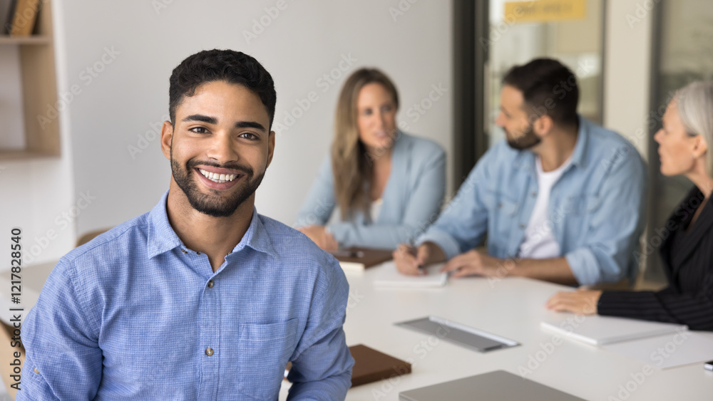 © fizkes - Young 20s Arabian male employee posing for camera seated at desk with colleagues on background, exudes competence, professionalism and reliability. Bank advisor, financial consultant portrait. Career