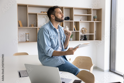 Pensive Hispanic employee working on creative ideas at modern workspace, deep in thoughts, holding pen and notepad, make notes, preparing insights for startuppers meeting, looks focused and thoughtful photo