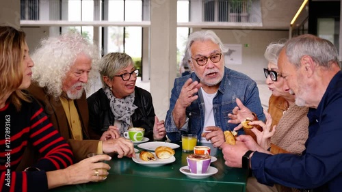 Group of joyful elderly friends gathered on terrace of residence cafeteria eating and drinking coffees and pastries. Caucasian seniors people meeting enjoying snack sitting together at table