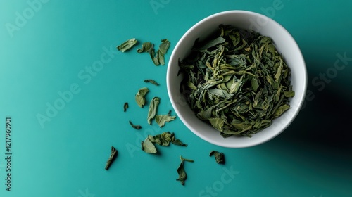 Scattered sencha green tea leaves in a white bowl against a vibrant teal background showcasing the beauty of natural tea