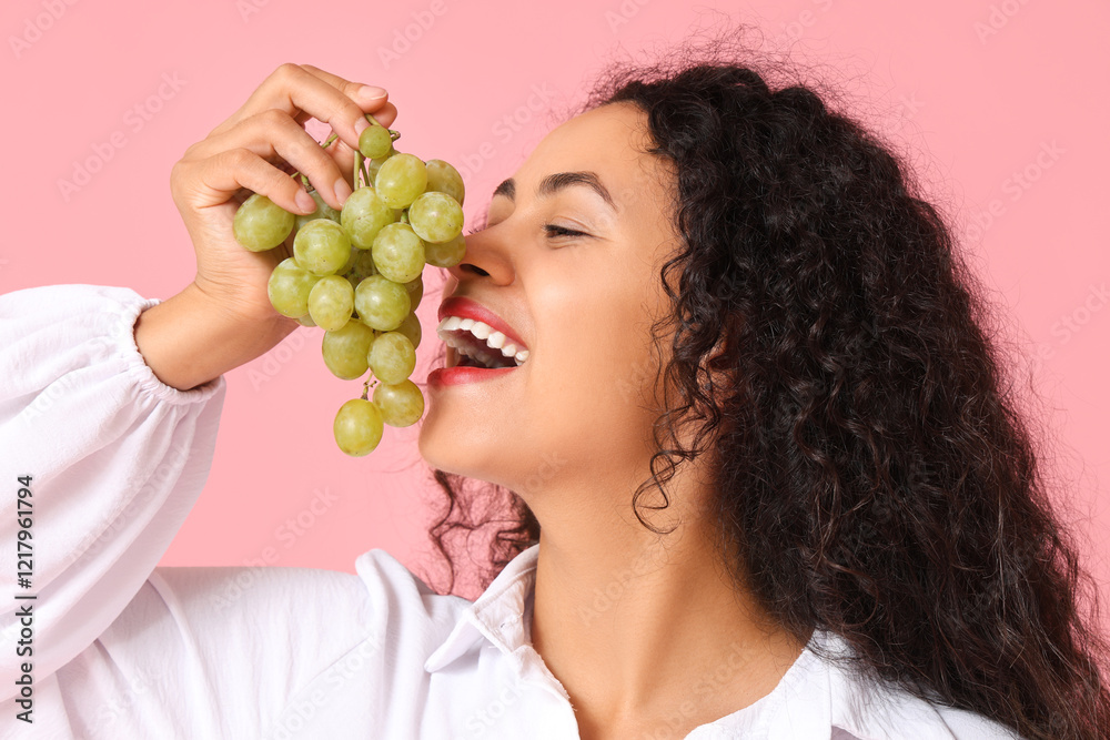 Beautiful young African-American woman eating sweet ripe grapes on pink background Beautiful young African-American woman eating sweet ripe grapes on pink background