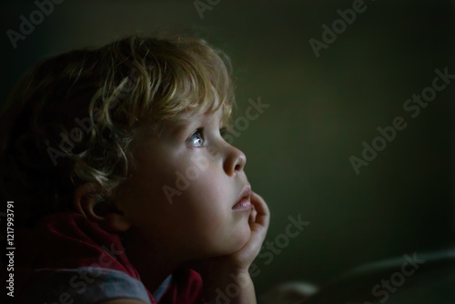 Young male Caucasian toddler boy laying in the dark looking up enthralled at a television tv or computer screen monitor