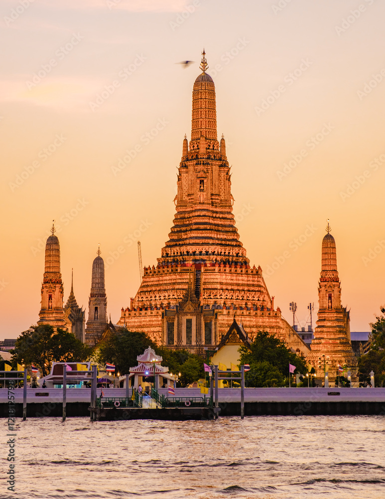 Fototapeta premium A captivating view of Wat Arun Bangkok as the sun sets, casting warm hues on the intricate temple. Visitors admire the beauty from the riverside, creating a peaceful ambiance as day turns to night.
