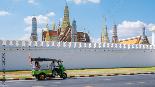 A colorful tuk-tuk whizzes past the grand walls of the iconic Grand Palace in Bangkok. The bright structure gleams in the daylight, surrounded by traditional architecture and ornate spires. photo