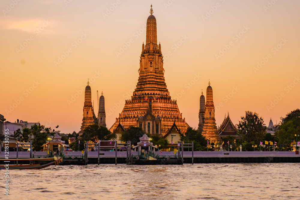 Fototapeta premium Golden light bathes Wat Arun, the Temple of Dawn, as dusk descends over Bangkok. The majestic spires reflect elegantly on the river, creating a serene atmosphere for evening strolls.